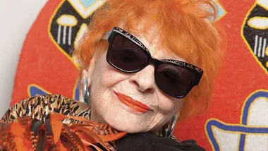 Senior woman with bright orange hair and stylish sunglasses, wearing patterned outfit against a vibrant backdrop.