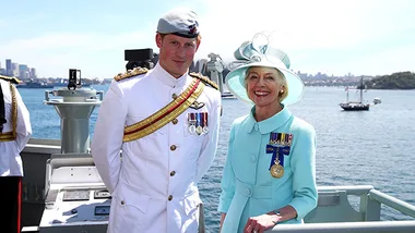 Two people in formal attire with military medals stand on a ship deck with water and city skyline in the background.