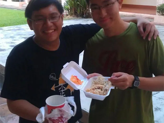 Two people smiling, one holding a drink and the other holding a takeout box with food, standing outside on a sunny day.