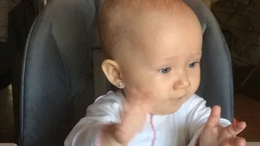 Baby sitting on a high chair, wearing a white outfit, with pierced ears.