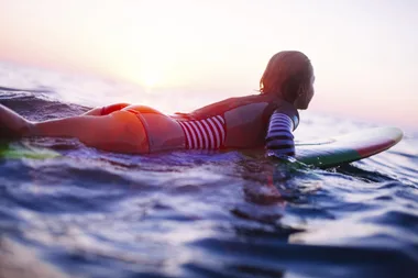 Teen surfer paddling on a board in the ocean at sunset, wearing a striped wetsuit.
