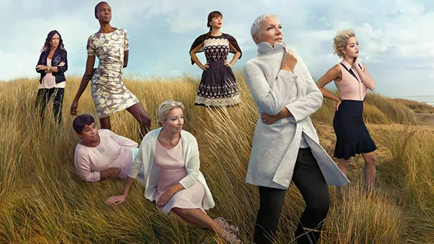 Seven women in stylish clothing pose in a grassy field, showcasing a diverse fashion campaign under a cloudy sky.