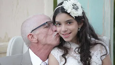 Elderly man in glasses kisses young girl in white dress with floral headband, both smiling warmly.