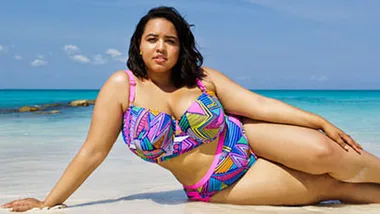 Plus-size woman in a colorful bikini lounging on a tropical beach with clear blue water and sky.