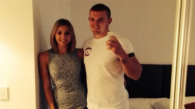 Couple taking a selfie in a bedroom, the man in a white t-shirt and the woman in a sleeveless gray dress.