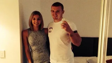 Couple taking a selfie in a bedroom, the man in a white t-shirt and the woman in a sleeveless gray dress.