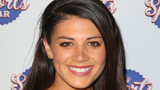 A woman with long dark hair and a bright smile poses against a sports-themed backdrop.