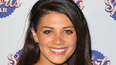 A woman with long dark hair and a bright smile poses against a sports-themed backdrop.