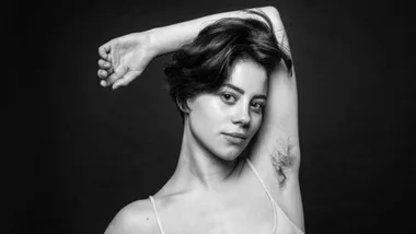 Portrait of a woman confidently showing natural armpit hair, against a dark background.