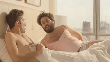 Two men in a bed, one with a beard wearing a pink camisole, the other shirtless, looking surprised.