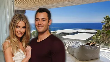 A couple smiles in front of a luxurious beachside property with ocean view in the background.