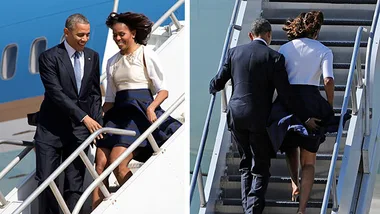 Michelle Obama and companion descend an airplane stairway, with wind causing her dress to billow.