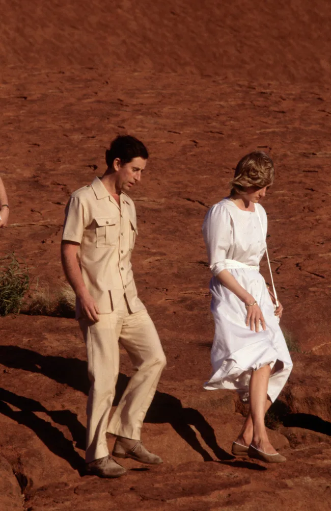 A couple walking down a rocky, reddish terrain; man in beige, woman in white dress.