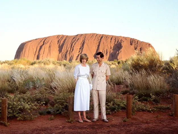 A man and woman stand in front of Uluru, Australia, with dry grass and clear sky in the background.