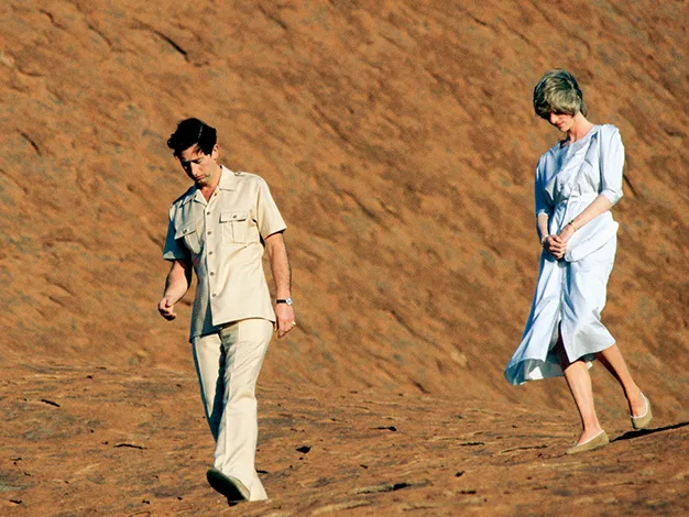 Two people walking on a red sandy surface; the man in a beige outfit, the woman in a white dress.