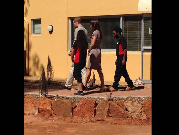 Four people walking outside a yellow building, two in red and black uniforms, with a stone wall in the foreground.