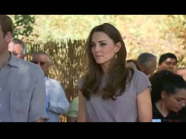 A woman with long brown hair stands outdoors, surrounded by people, under a sunny sky.