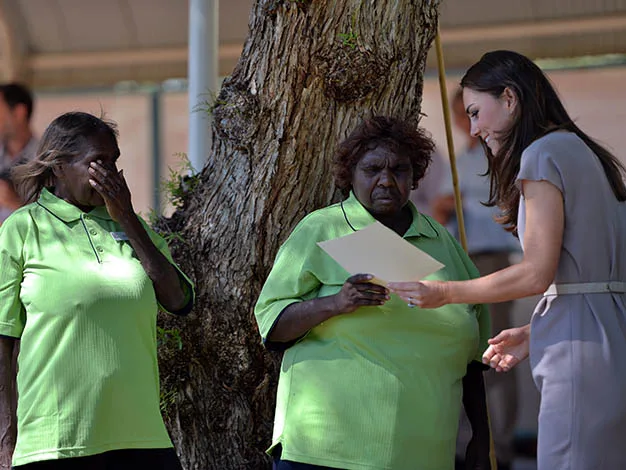 Two women in green shirts and a woman in a gray dress stand by a tree, looking at a paper together.