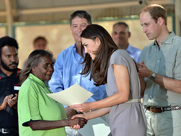 A woman in a beige dress shakes hands with an elderly woman holding a certificate, while others look on.