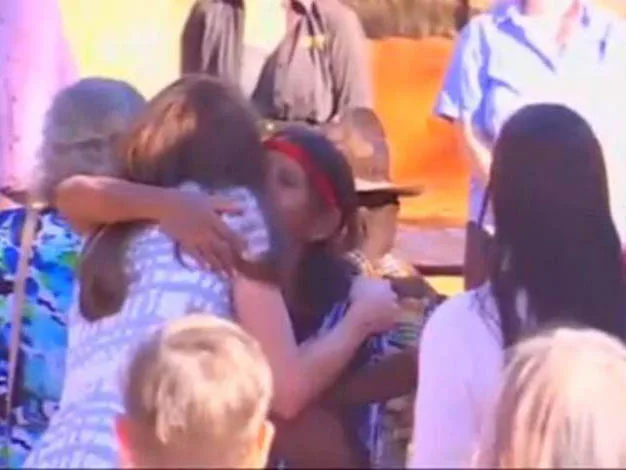 A group of people gathered outdoors, one woman hugging another, with others watching under a sunny sky.