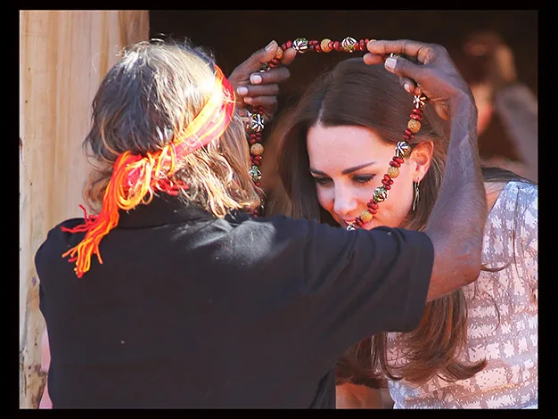 A person places a beaded necklace over a woman's head. The woman is leaning slightly forward, with a small smile.