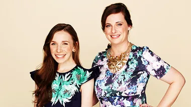 Two women smiling, wearing vibrant floral-patterned dresses, standing against a beige background.