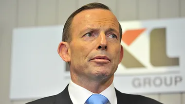 A man in a suit and blue tie stands before a blurred background sign with partial lettering visible.