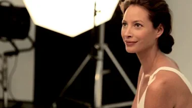 Christy Turlington smiling at a photoshoot, wearing a white top, with studio lights in the background.