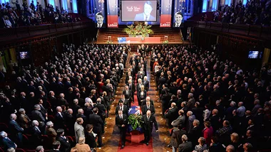 State funeral for Neville Wran, with attendees gathered, portrait on screen, and flag-draped casket in the center aisle.