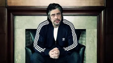 Man in Adidas tracksuit sitting in a chair, hands clasped, speaks directly to camera in a serious tone.