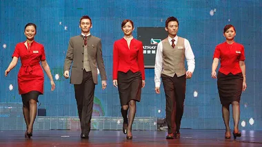 Flight attendants walking in uniform on a stage; women in red, men in gray and white, with a logo in the background.