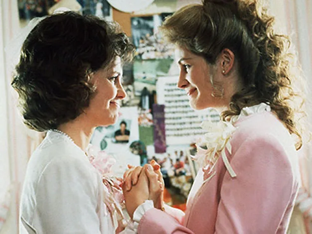 Two women smiling, holding hands in a warmly lit room, wearing pastel outfits with floral corsages.