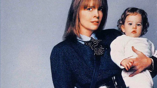 A woman in a blue suit holds a baby in white clothing against a plain background.