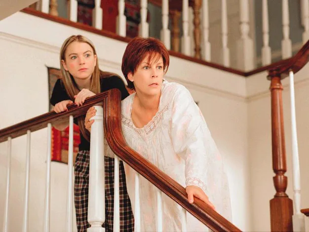 Two women, one in pajamas leaning on a staircase railing, looking surprised inside a house.
