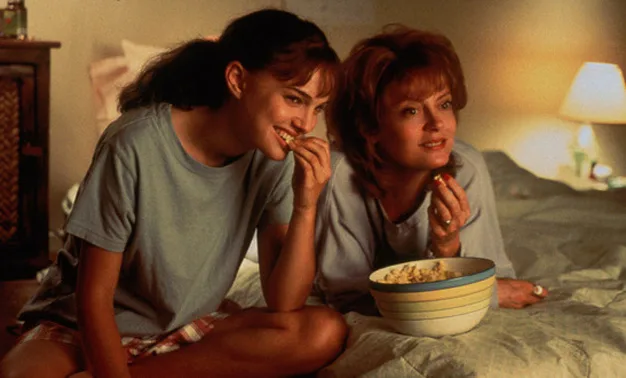 Two women sit on a bed, smiling and eating popcorn, suggesting a relaxed, cozy setting.