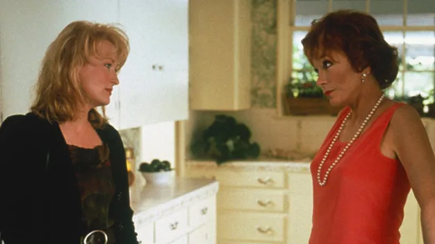 Two women stand in a kitchen facing each other, one in a black jacket and the other in a red top with pearls.