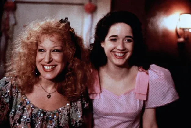 Two women smiling, one with curly blonde hair in a patterned dress and heart necklace, the other with dark curly hair in a pink dress.