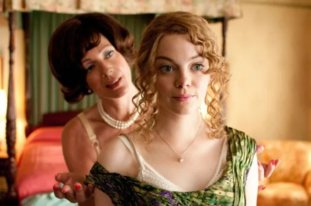 Two women in a bedroom; the one in the background adjusts the other's dress. Both appear thoughtful.
