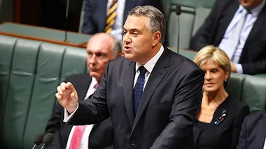 A man in a suit speaks passionately at a government assembly, with colleagues seated around him.