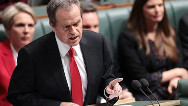 Leader giving a speech in parliament, gesturing with hand, with colleagues seated behind.