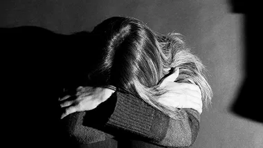 Person sitting with their head down, embracing their arms, in a dimly lit environment conveying a sense of distress.