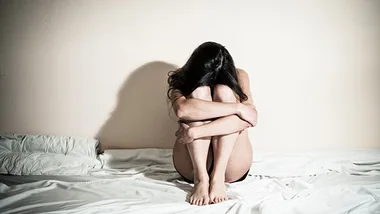 A woman sitting on a bed, hugging her knees, with her face hidden in her arms.