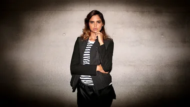 A woman in a striped shirt and black jacket stands confidently against a textured wall, with a thoughtful expression.