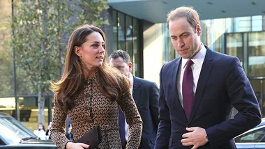 A man and woman walking together, dressed formally in suits, outside a modern glass building.