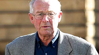 Man in glasses and jacket outdoors with blurred background.