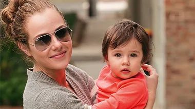 A woman in sunglasses holds a toddler in her arms outdoors, both looking at the camera.