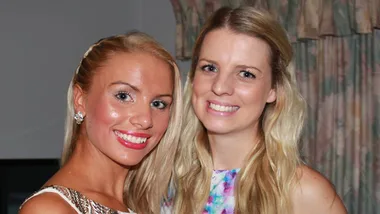 Two women smiling, one in a white dress with sparkly detail, the other in a floral dress, standing indoors.
