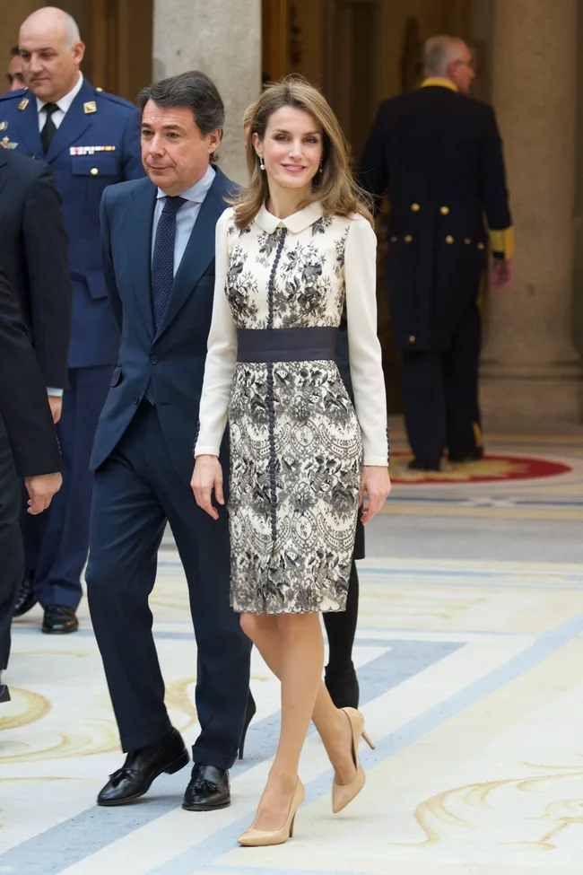 A woman in a patterned dress and beige heels walks with a man in a suit in a formal setting.