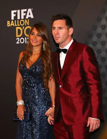Ballon d'Or 2013 event: A couple poses on the red carpet. The man wears a red suit, the woman a navy blue gown.