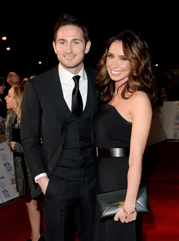 Man in a suit and woman in a black dress smiling on a red carpet event, with people in the background.